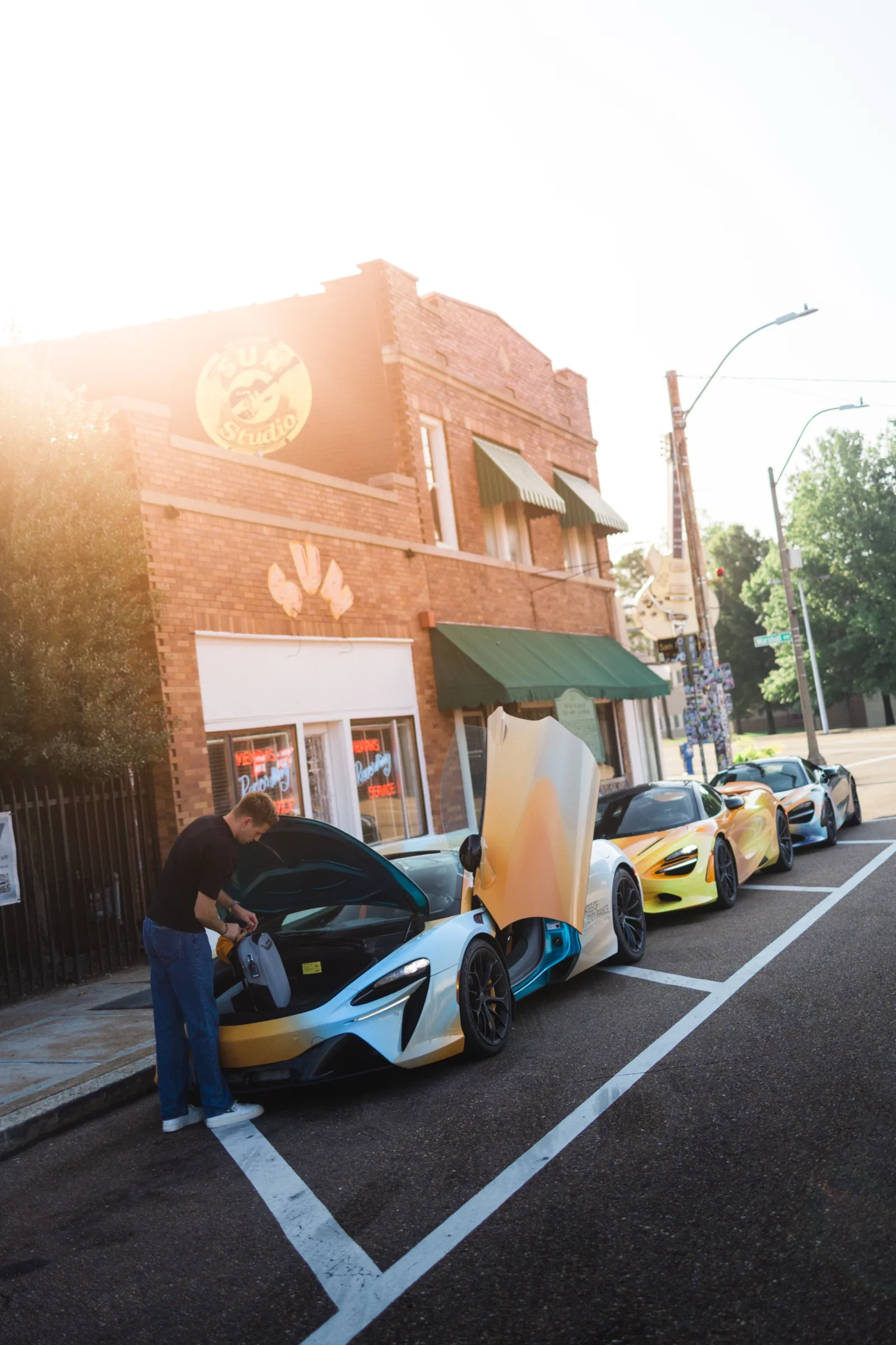 Jack loading luggage into a McLaren outside Sun Studio in Memphis at sunrise, three McLarens lined up along the kerb